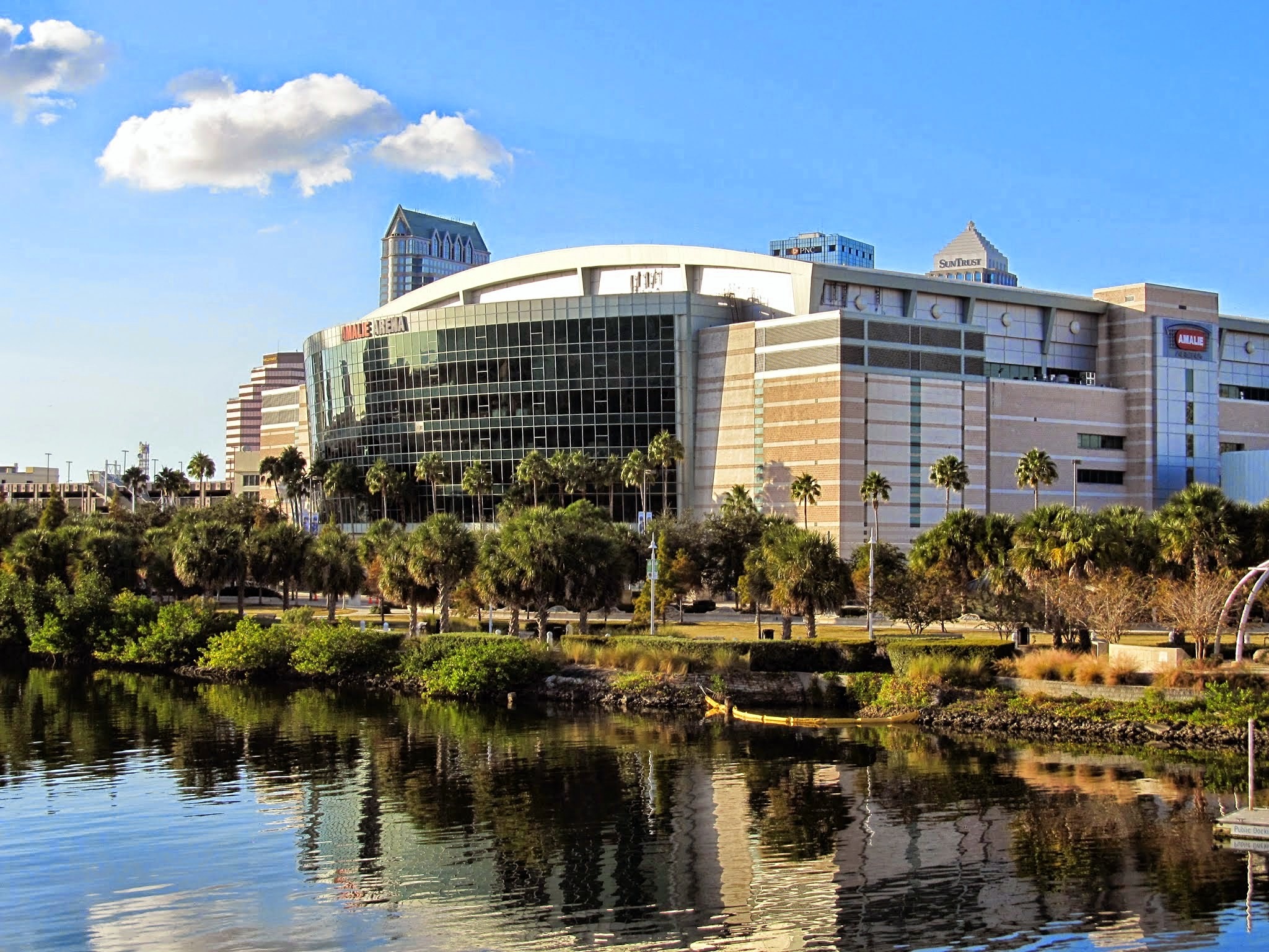 Benchmark International Arena viewed from across the Tampa Riverwalk waterway, palm trees in foreground