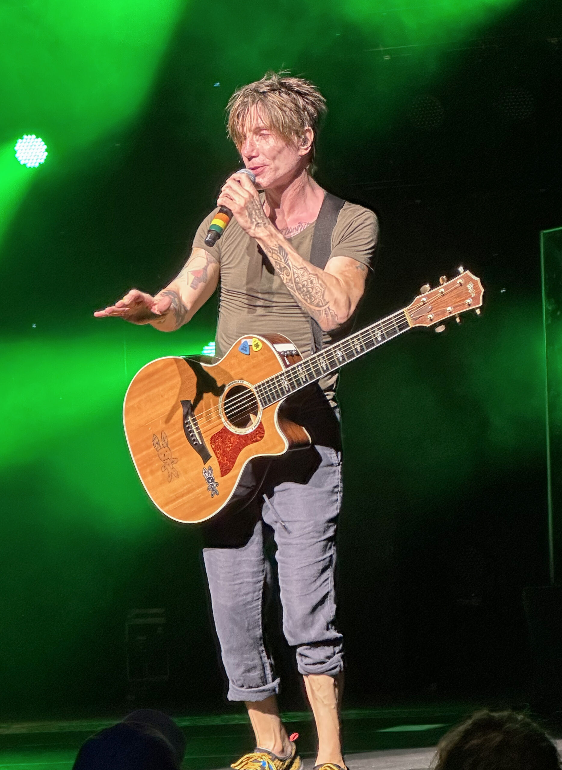 John Rzeznik of the Goo Goo Dolls performing live on stage with an acoustic guitar under green stage lighting — photo from a 2023 show in Syracuse, New York, not the upcoming Tampa date.