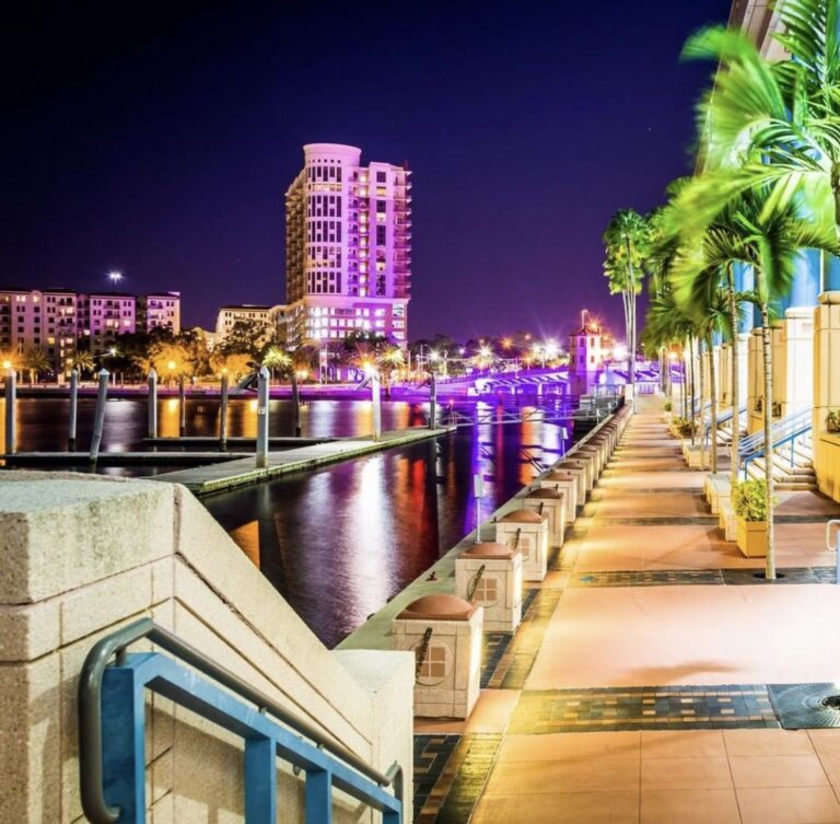 A nighttime view along the Tampa Riverwalk near the Tampa Convention Center, with palm trees lining a lit walkway beside the Hillsborough River and a purple-lit high-rise in the background.